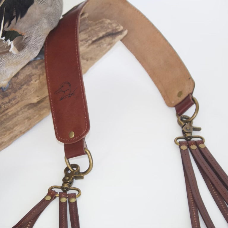 A close-up of a brown leather duck hunting strap with brass hardware and multiple hanging loops, displayed against a white background with a taxidermy duck and wood prop.