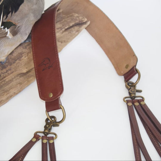 A close-up of a brown leather duck hunting strap with brass hardware and multiple hanging loops, displayed against a white background with a taxidermy duck and wood prop.