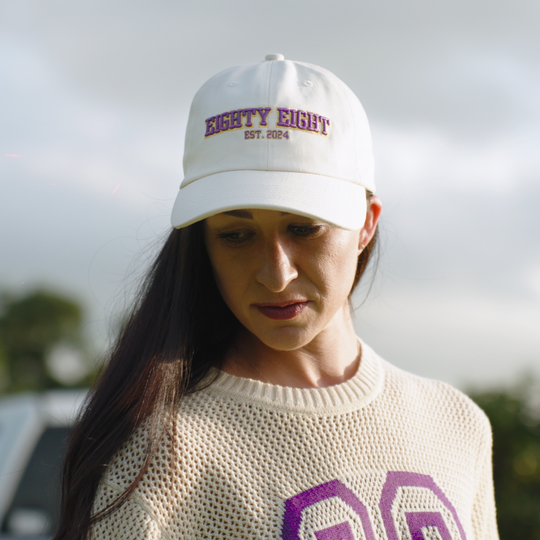 A woman wearing a white baseball cap with purple and maroon embroidered text reading &