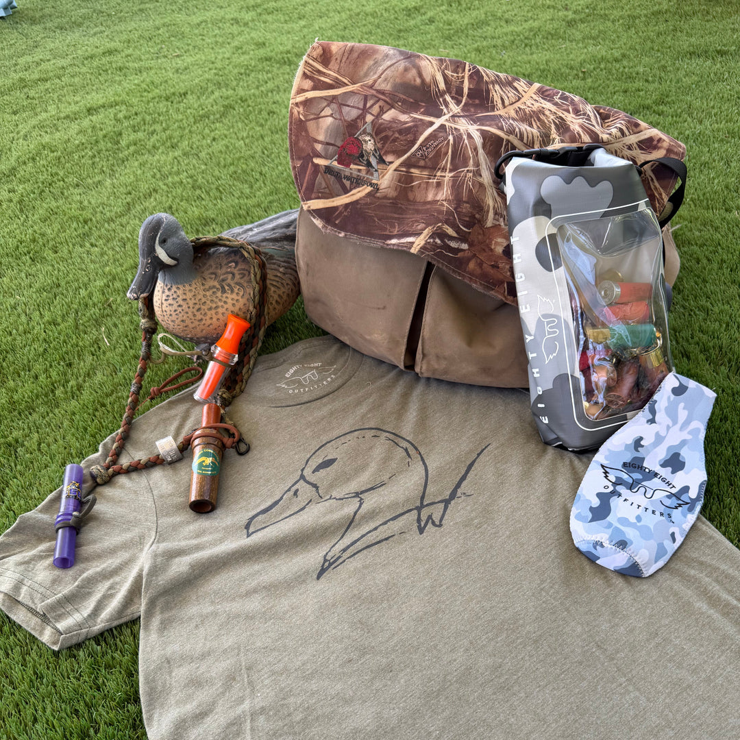 A flat-lay of a duck hunting merchandise set on green grass, featuring a heather olive green t-shirt with a black duck outline, a camouflage duck blind, a camouflage-patterned bag, a duck deco, a bow, and a pack of fishing lures.