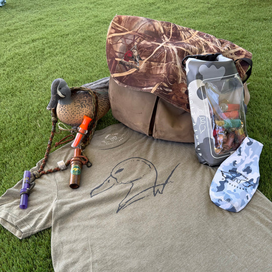 A flat-lay of a duck hunting merchandise set on green grass, featuring a heather olive green t-shirt with a black duck outline, a camouflage duck blind, a camouflage-patterned bag, a duck deco, a bow, and a pack of fishing lures.