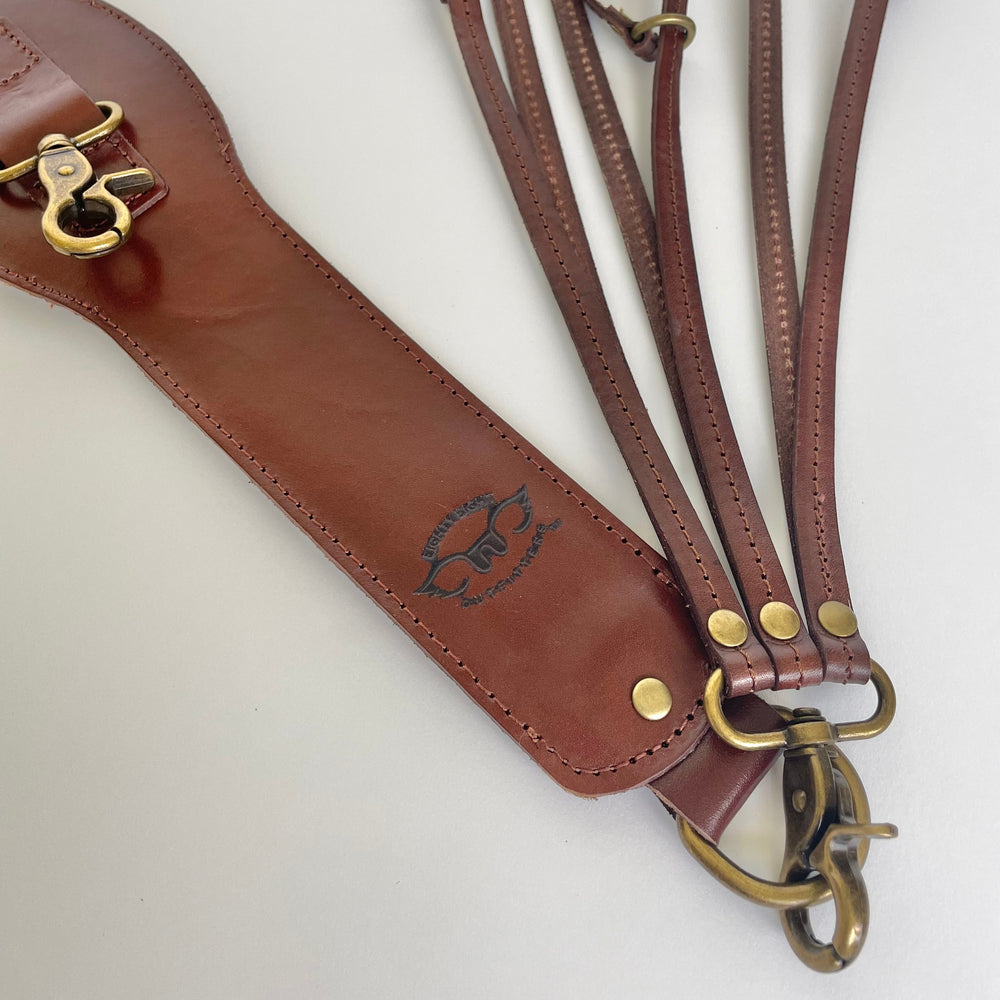 A brown leather strap set with brass hardware, laid out on a light off-white background. The set includes a main strap with a snap hook and a keeper loop