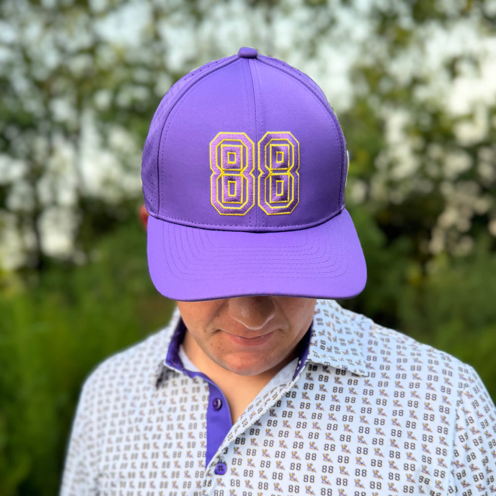 A man wearing a purple baseball cap with a gold and white embroidered '88' on the front, paired with a white patterned polo shirt, set against a blurred outdoor background of green foliage and white blossoms.
