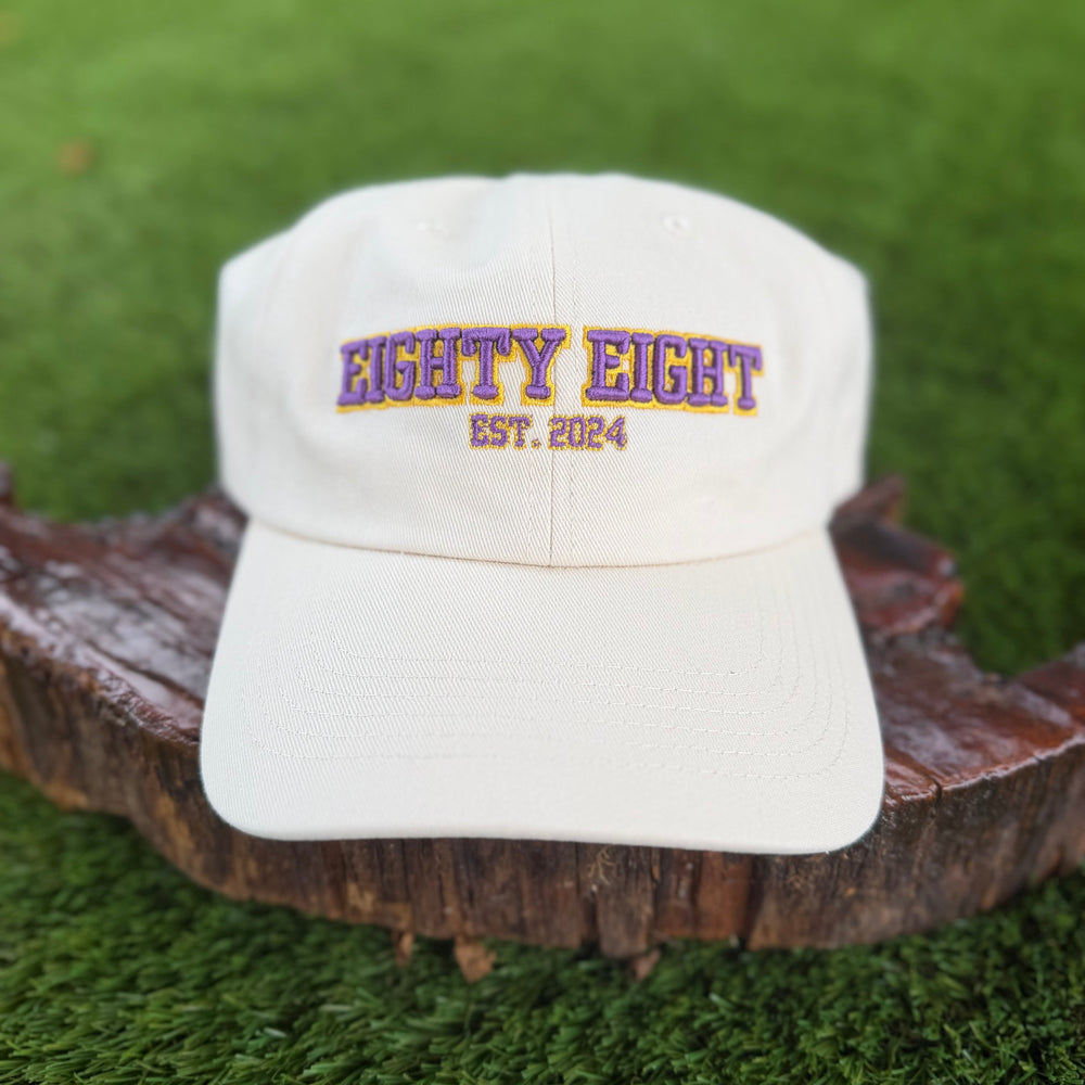 An off-white baseball cap with 'EIGHTY EIGHT EST. 2024' embroidered on the front, displayed on a rustic wooden log slice against a green grass background.