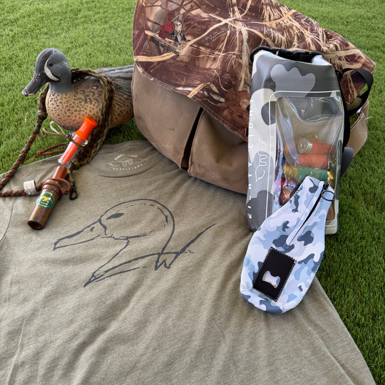A duck hunting apparel and gear flat lay on green grass, featuring a heather olive t-shirt with a duck outline, a camouflage duck bag, a camouflage sock, a deco duck, and a whistle.