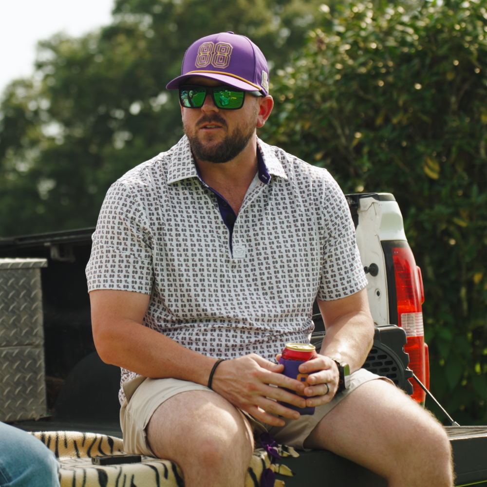 A man wearing a purple baseball cap with '88' on it, green-tinted sunglasses, a patterned short-sleeve polo shirt, and khaki shorts sits in the back of a pickup truck holding a can of beer.