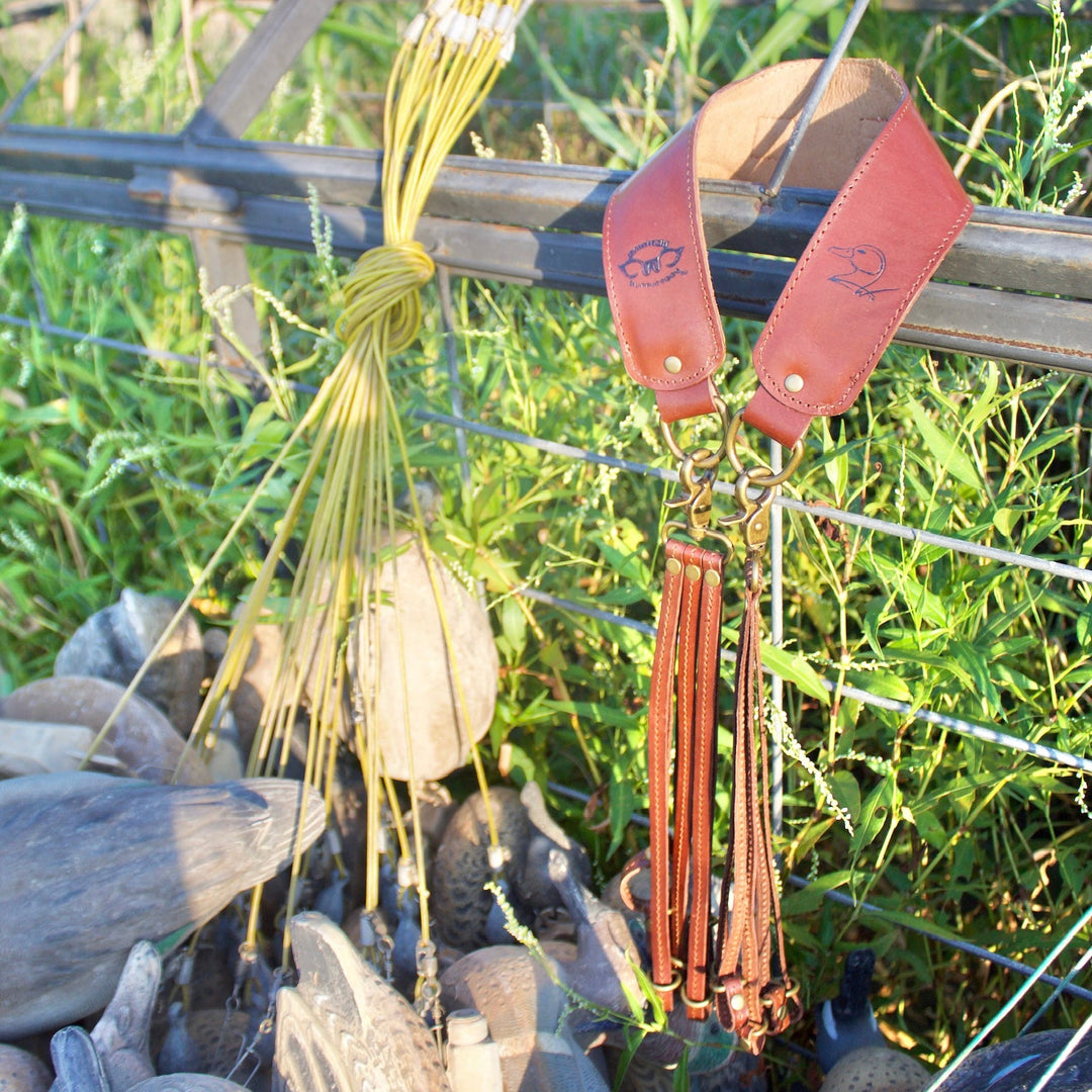 A set of duck hunting accessories hanging from a wire fence in an outdoor marsh. The set includes a bundle of yellow synthetic duck calls, a pair of reddish-brown leather quill bands with tassel ends, and a cluster of realistic duck deco ducks on the ground below.