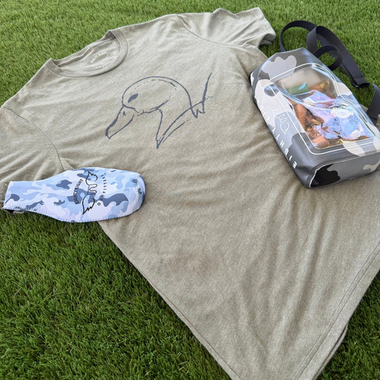 A heather olive green t-shirt with a simple line drawing of a dog, laid flat on green grass alongside a blue and white camouflage sock and a clear plastic bag.