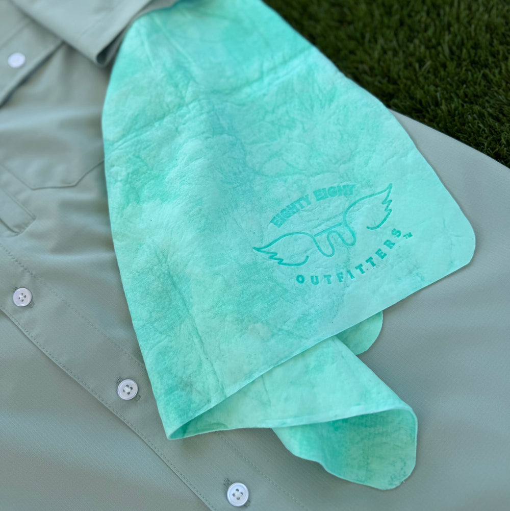 A mint green tie-dye bandana with a winged logo and the words 'Eighty eight OUTFITTERS' is draped over a light sage green button-up shirt, placed on green grass.