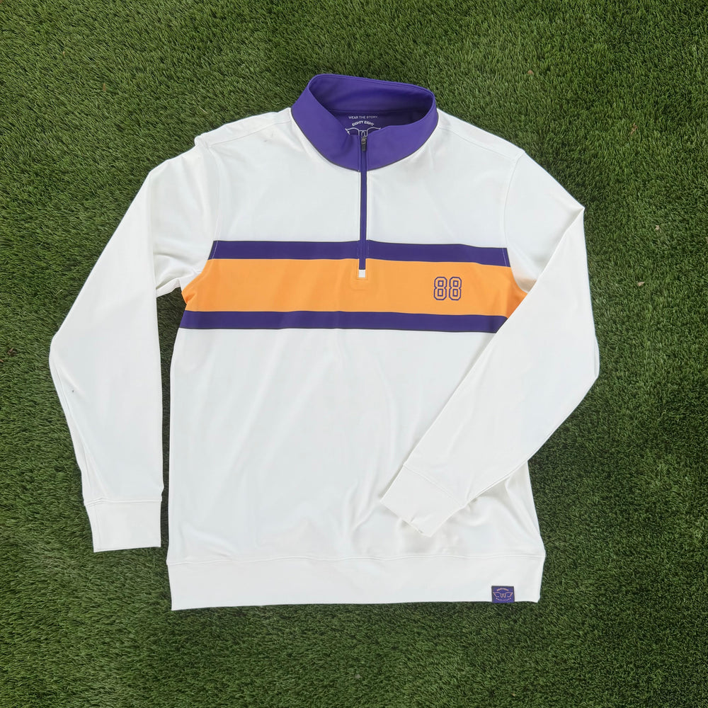 A white long-sleeve quarter-zip pullover with a purple collar and a horizontal orange and purple chest stripe, laid flat on green artificial turf.