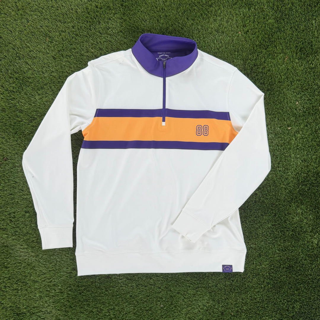A white long-sleeve quarter-zip pullover with a purple collar and a horizontal orange and purple chest stripe, laid flat on green artificial turf.