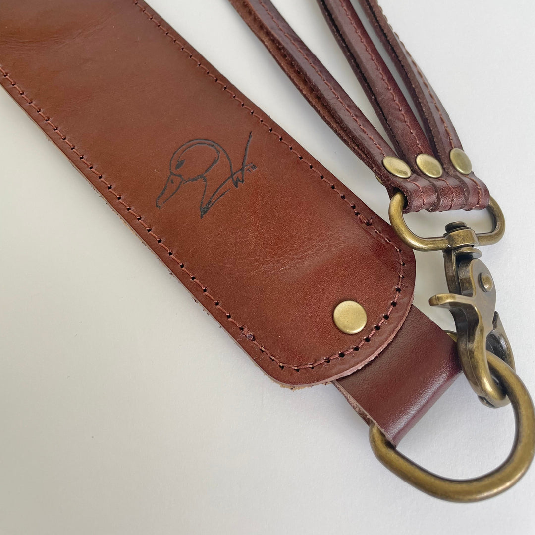 A close-up of a brown leather strap with brass hardware, including a snap hook, studs, and a large D-ring, laid on a light background. A small black duck sketch logo is embossed on the leather.