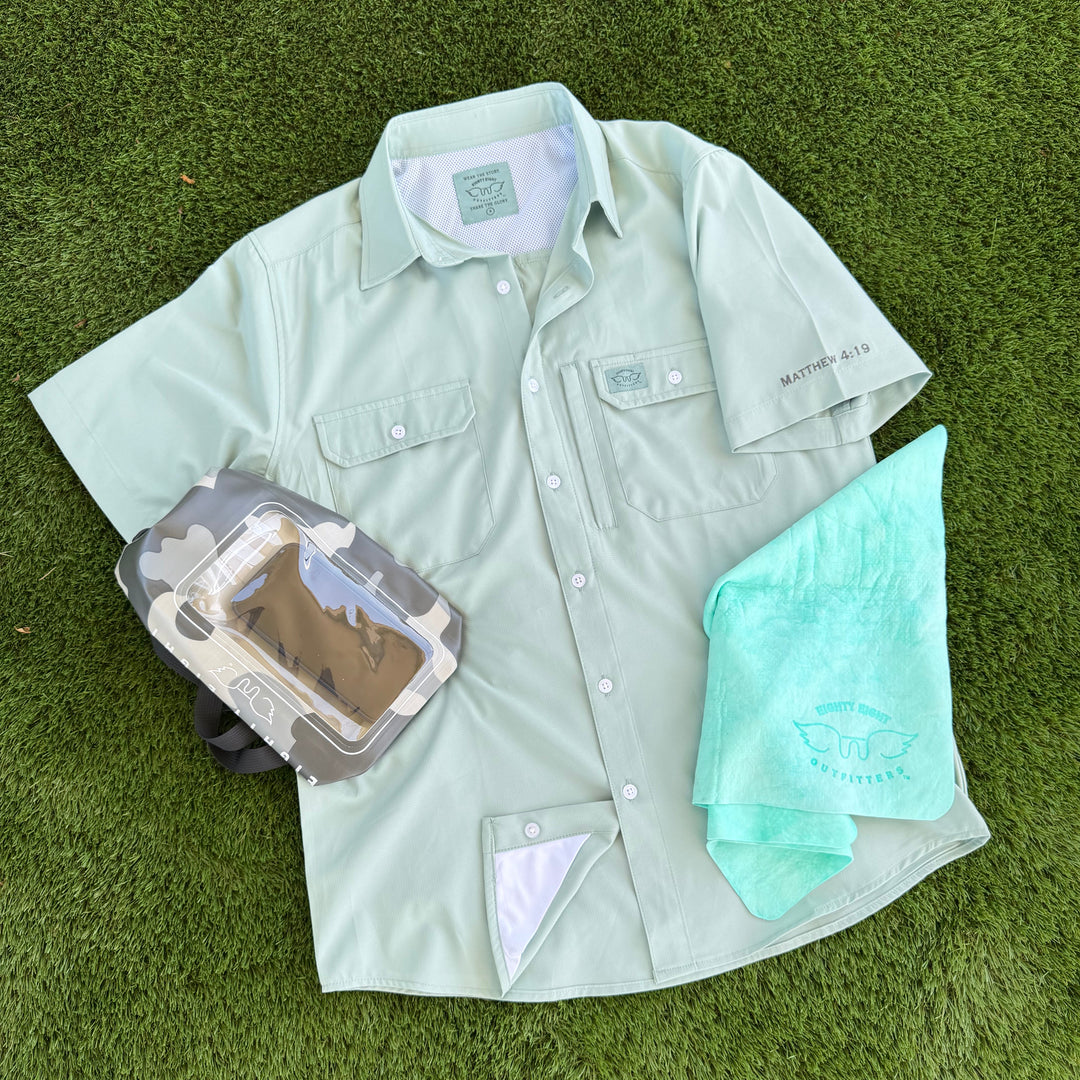 A light sage green short-sleeve button-up shirt laid flat on green grass, styled with a gray camouflage camera pouch and a mint green towel. The shirt features two chest pockets, a collar label, and a small script logo on the left sleeve.