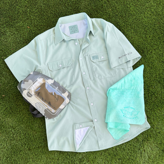 A light sage green short-sleeve button-up shirt laid flat on green grass, styled with a gray camouflage camera pouch and a mint green towel. The shirt features two chest pockets, a collar label, and a small script logo on the left sleeve.