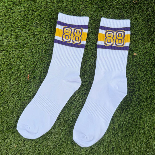 A pair of white crew socks with purple and yellow stripes and the number 88 on the cuff, laid flat on green grass.