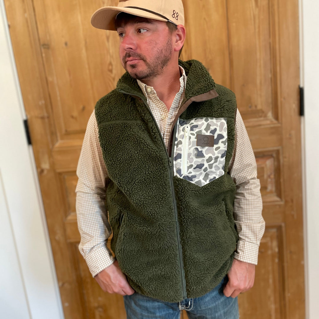 Man wearing a green vest with a patterned pocket, beige cap, and plaid shirt in front of wooden paneling.