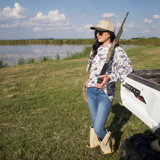 Woman standing by a truck bed with a rifle, near a body of water and grassy area.
