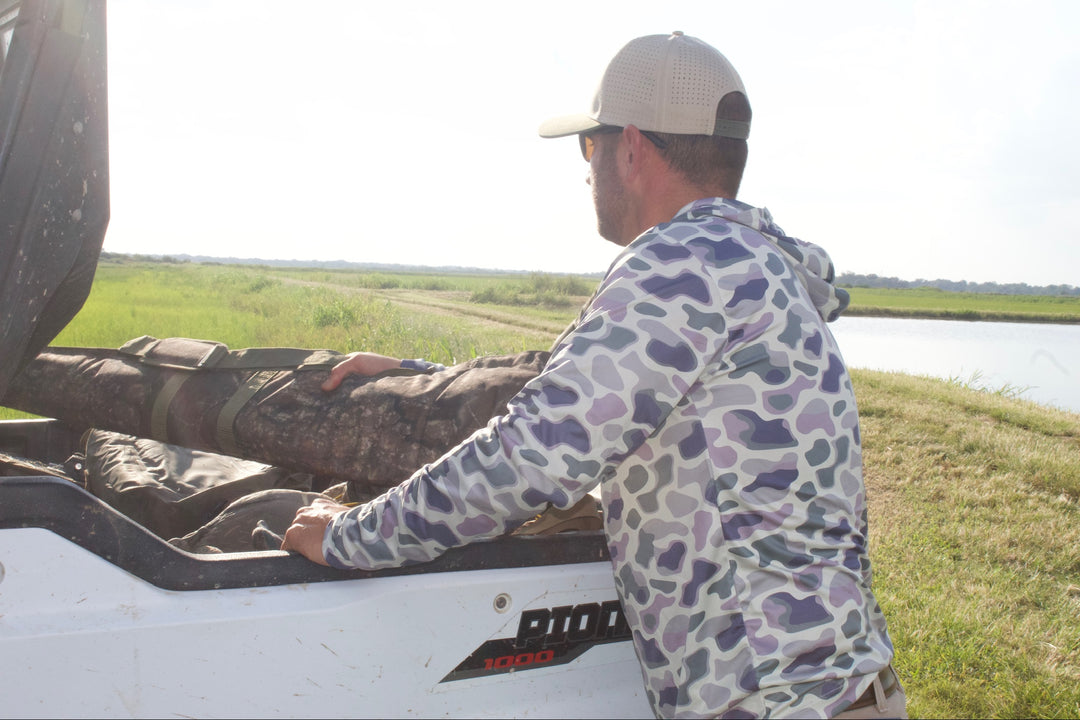 Man in camouflage jacket and cap opening a vehicle door in a grassy field with a lake in the background