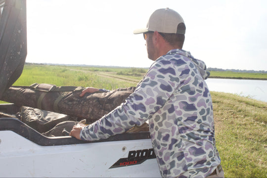 Man in camouflage jacket and cap opening a vehicle door in a grassy field with a lake in the background