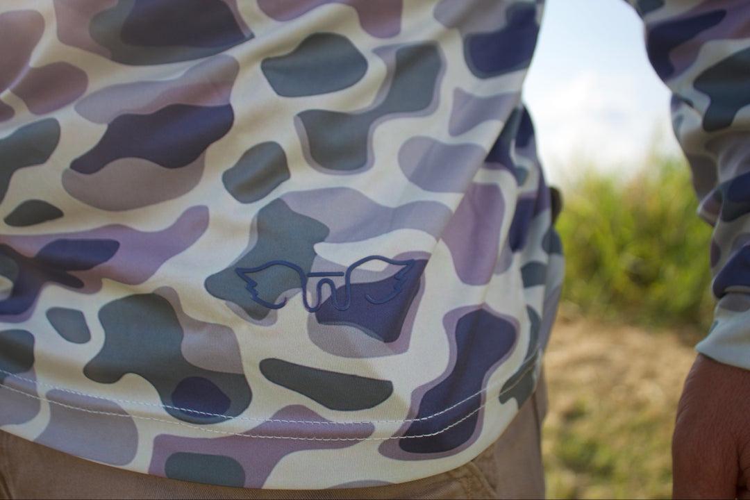Person wearing a camouflage-patterned shirt with a logo in an outdoor setting