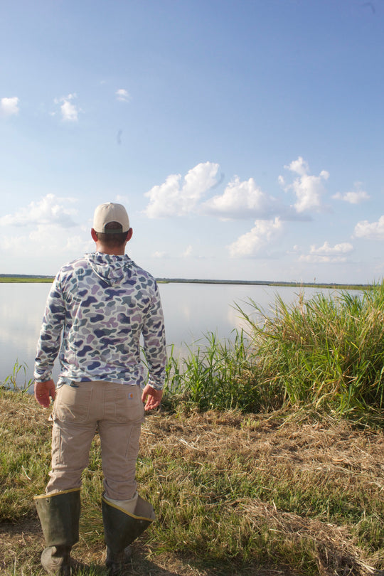 Person standing by a lake, wearing a patterned shirt and beige pants, with a blue sky and clouds in the background.