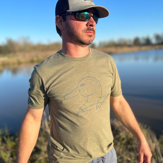 Man wearing a green t-shirt with a duck design, gray cap, and sunglasses, standing by a body of water.