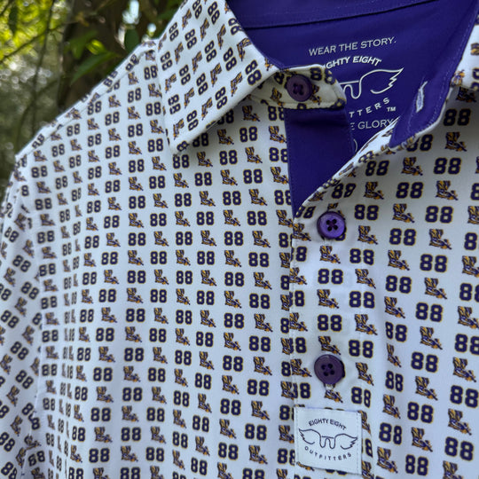 A close-up of a patterned collared shirt hanging on a black hanger outdoors. The shirt has a white base with an all-over repeating dark brown and navy graphic pattern, a solid navy collar, and matching navy buttons.