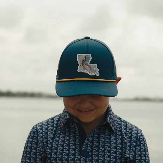 Child wearing a blue cap with a pelican design, standing by a body of water.