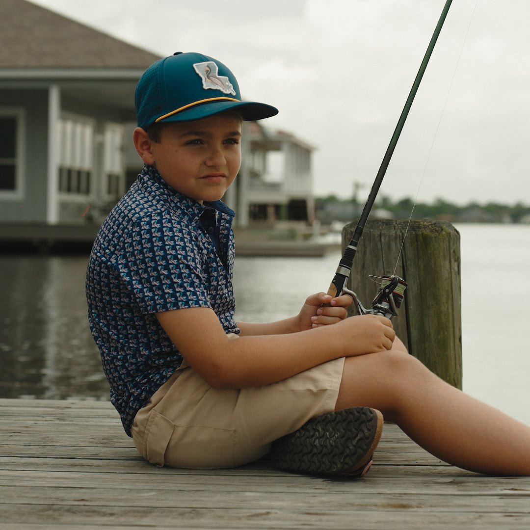 Child sitting on a dock with a fishing rod, wearing a blue cap and patterned shirt.