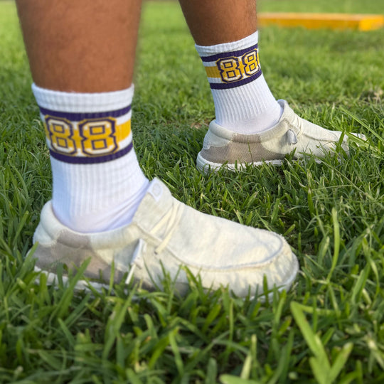 White athletic crew socks with yellow and navy blue stripes and the number 88, worn with white slip-on shoes on a green grass field.