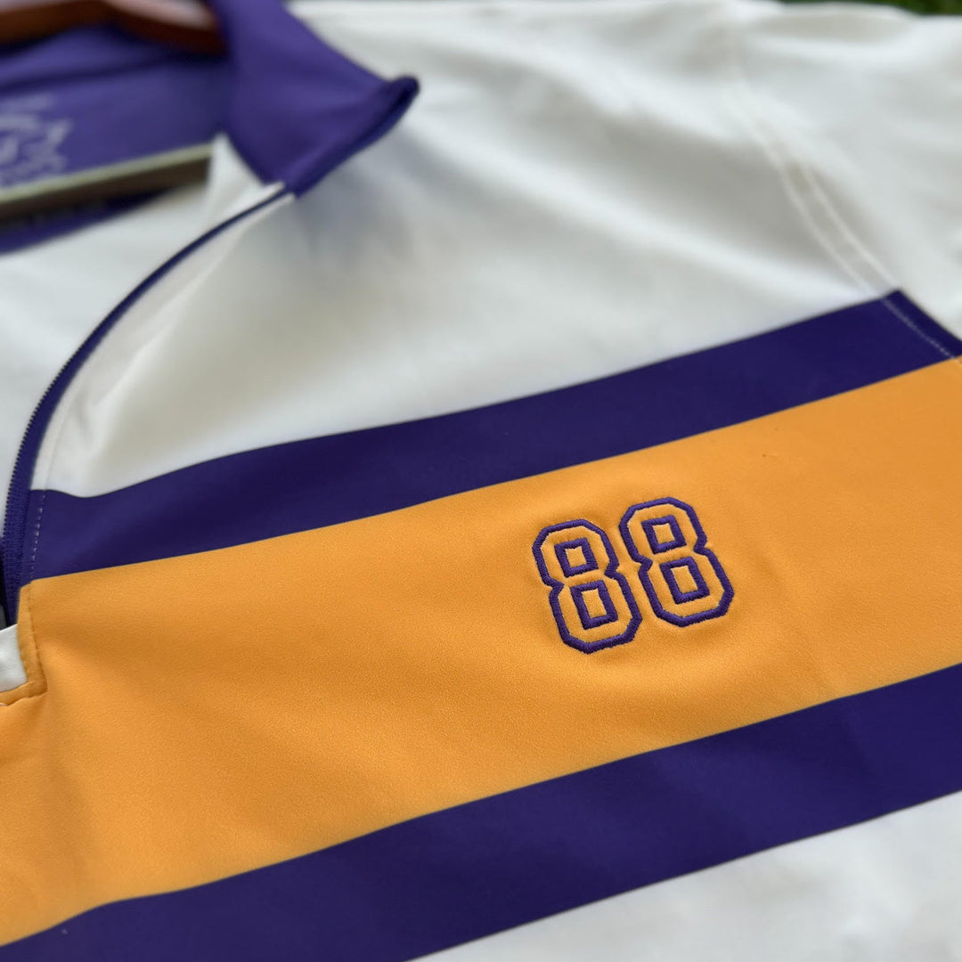 A close-up of a white athletic jersey with a wide yellow stripe across the chest, bordered by purple stripes, featuring the embroidered number 88 in purple outlined in yellow. The collar is purple, and the jersey is laid flat on green grass.