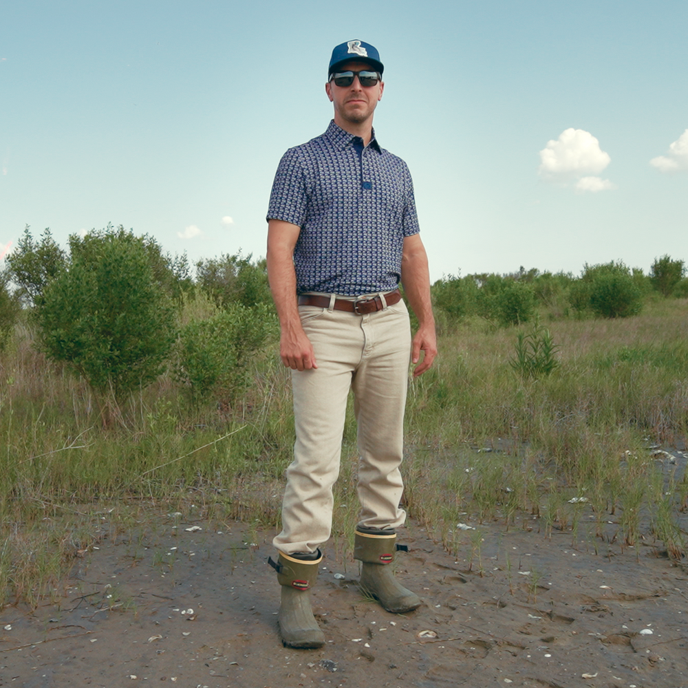 Man standing in a natural setting wearing a blue shirt, beige pants, and green boots.