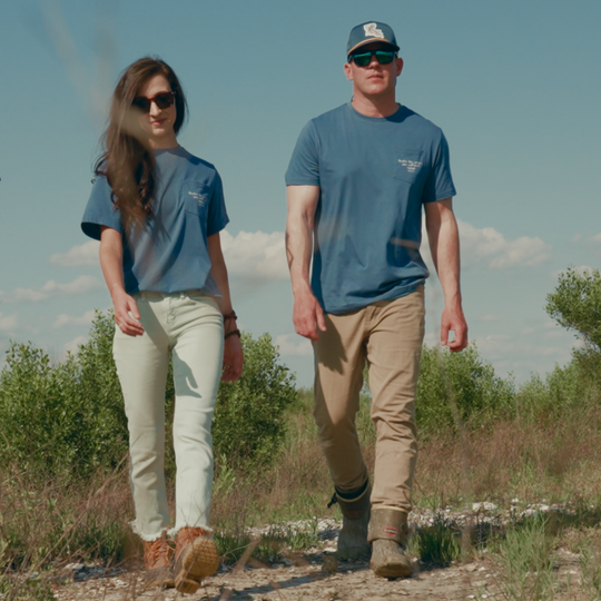 Two people standing on a dirt path with a clear sky and greenery in the background