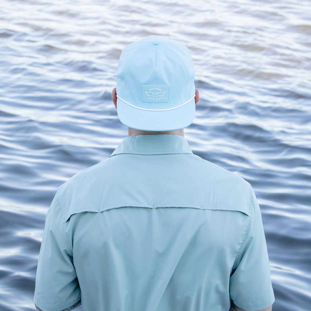 Person wearing a light blue cap and shirt standing by a body of water.