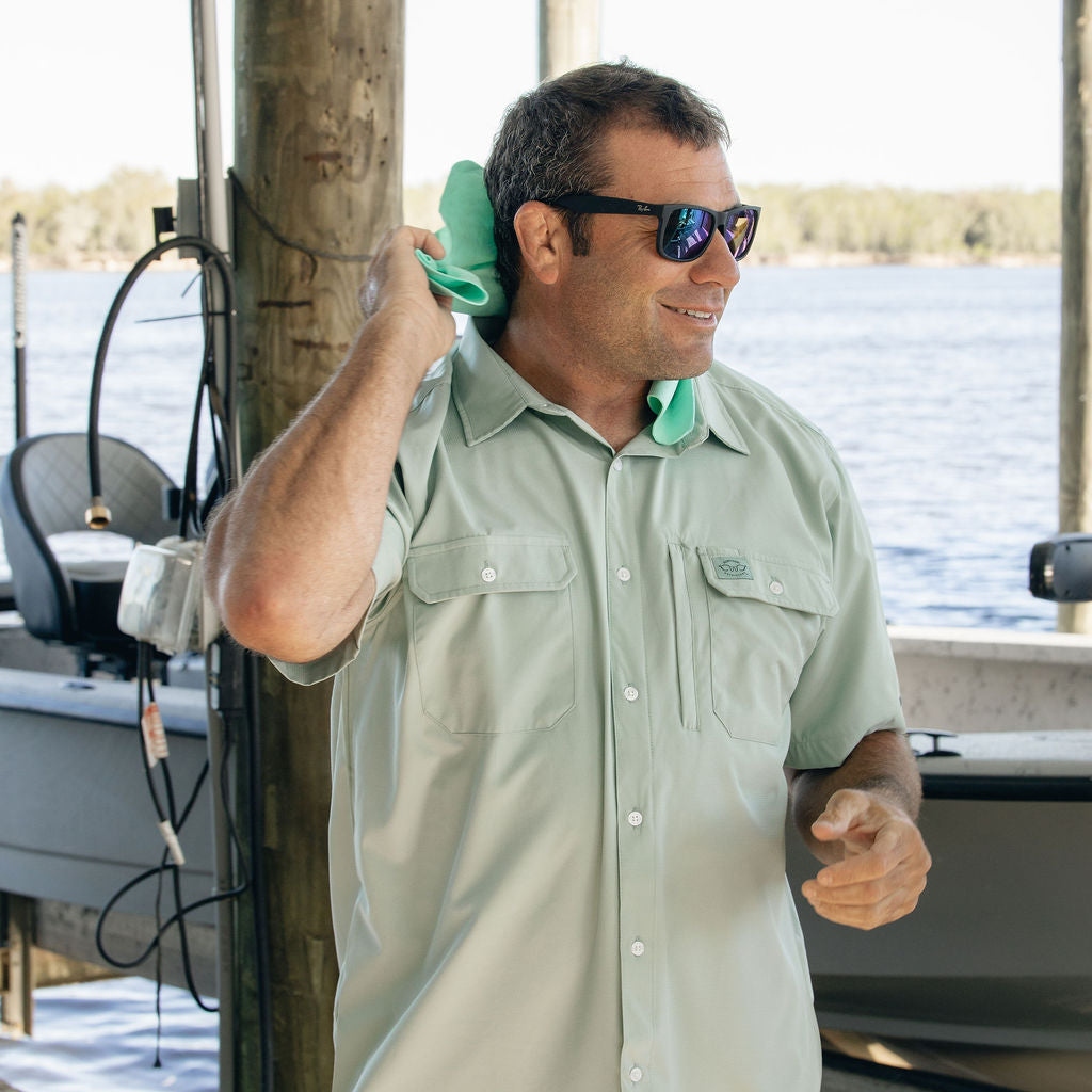 Man wearing sunglasses and a light green shirt by a lake with a boat in the background