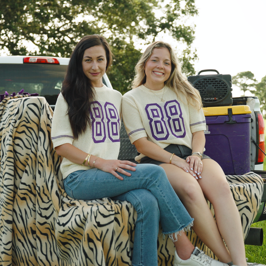 Two women sitting together outdoors in front of a pickup truck, wearing matching cream ribbed short-sleeve shirts with large purple number 88 graphics on the front. One is draped over a tiger-print blanket, and coolers and a speaker are visible in the background.